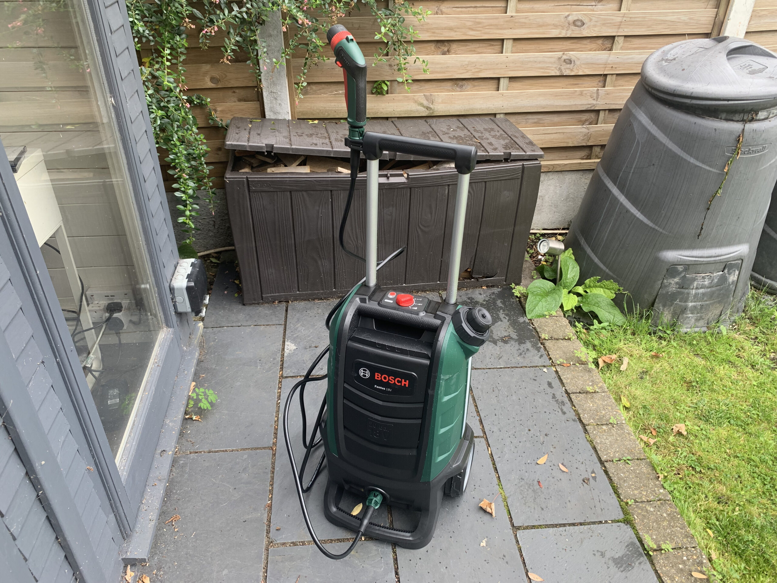 Bosch Fontus 18V sobre un patio exterior, junto a un tonel de agua y un baúl de madera, con el logo Bosch visible
