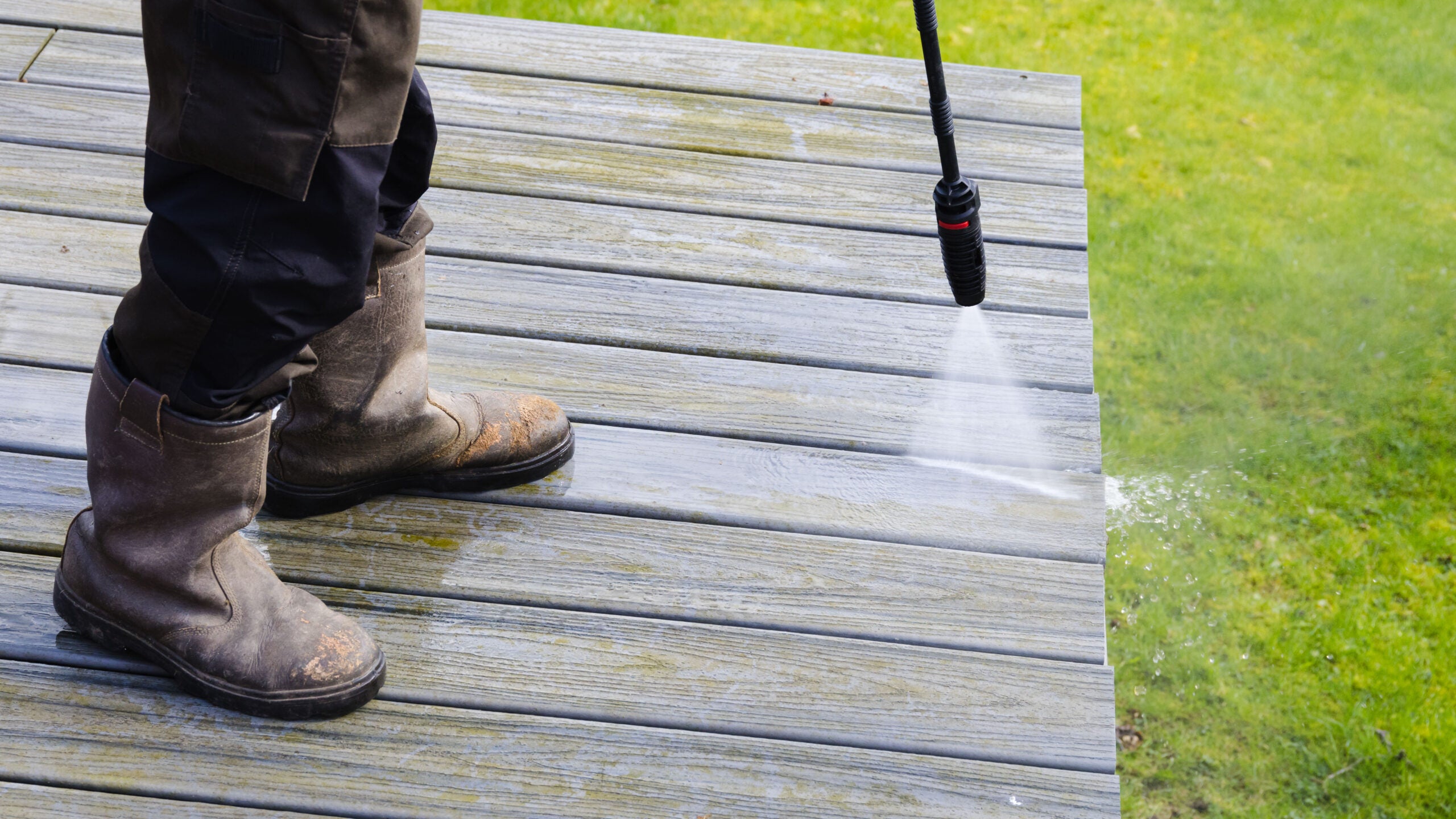 Persona limpiando una terraza de madera con hidrolimpiadora de alta presión, agua saliendo en abanico sobre la madera
